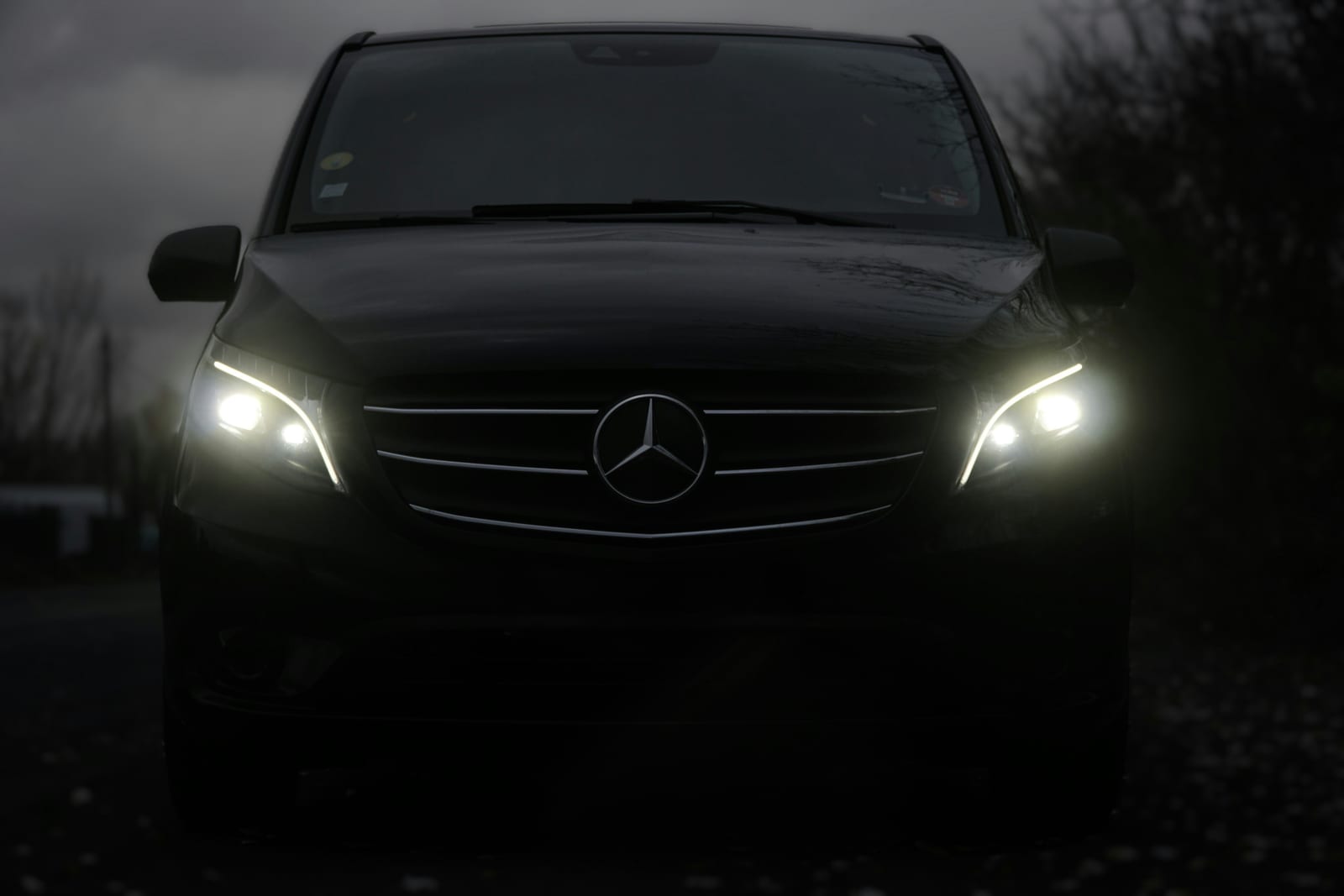 Van Mercedes-Benz preta, vista de frente, com os faróis acesos ao anoitecer